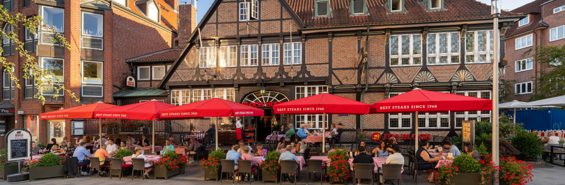 Außenansicht des BLOCK HOUSE Hamburg Bergedorf mit gut besuchter Terrasse, roten Sonnenschirmen und historischem Fachwerkgebäude.
