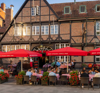 Außenansicht des BLOCK HOUSE Hamburg Bergedorf mit gut besuchter Terrasse, roten Sonnenschirmen und historischem Fachwerkgebäude.