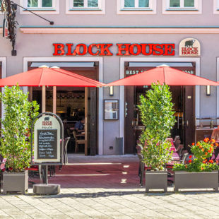 Außenansicht des BLOCK HOUSE Restaurants in Augsburg: eine große, hölzerne Fensterfront, davor viele Sitzgelegenheiten auf der Terrasse, die sich direkt auf dem Ulrichsplatz befindet.