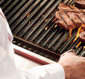 Blick über die Schulter eines Grillers, der eine Steakzange in der Hand hält und gerade ein Steak auf einem flammenden Grill wendet.