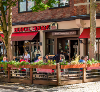 Außenansicht des BLOCK HOUSE Kiel mit Terrasse, roten Markisen und Gästen vor dem Restaurant.